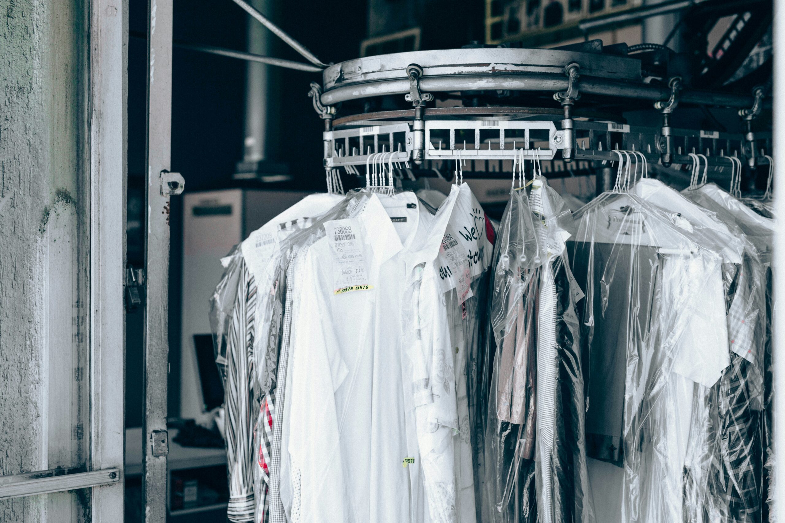 Clothes covered in plastic hang on a rotating rack inside a dry cleaning shop, with some tags visible on the garments and industrial dry cleaning equipment in the background.