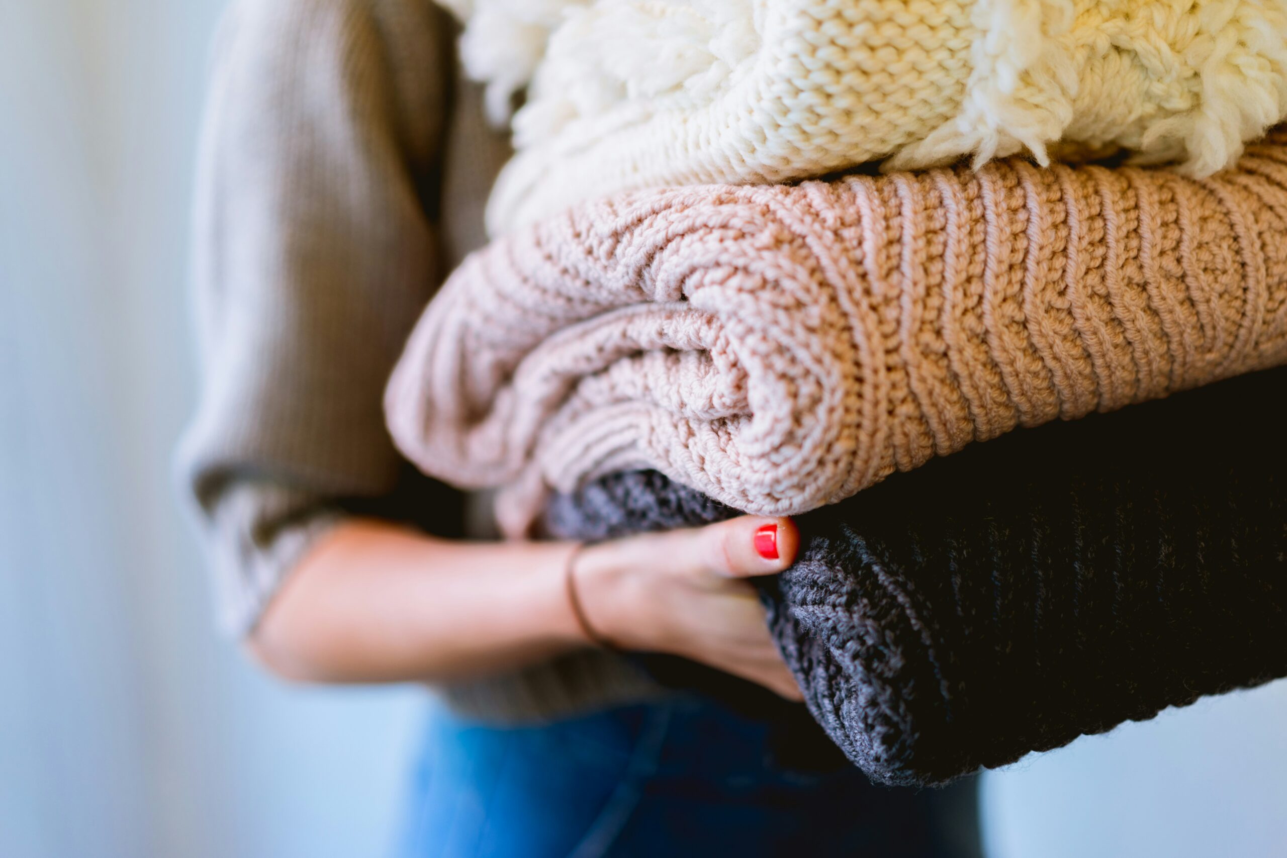 A person with red nail polish holds a stack of folded knitted sweaters in cream, light brown, and dark gray—showcasing how to make your clothes last longer. The persons face is not visible, and the softly blurred background adds warmth.