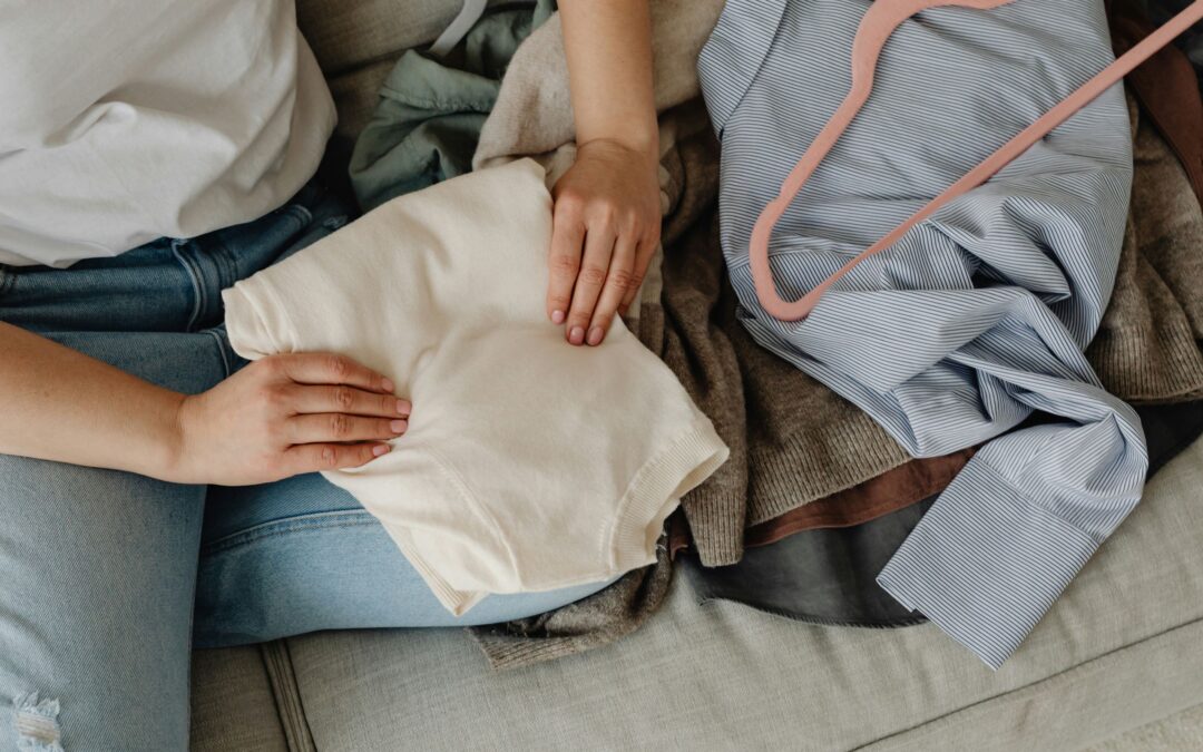 A person sitting on a couch wears jeans and a white shirt while folding a cream-colored garment. Other clothes, a pink hanger, and diy stain removal supplies are spread out beside them.