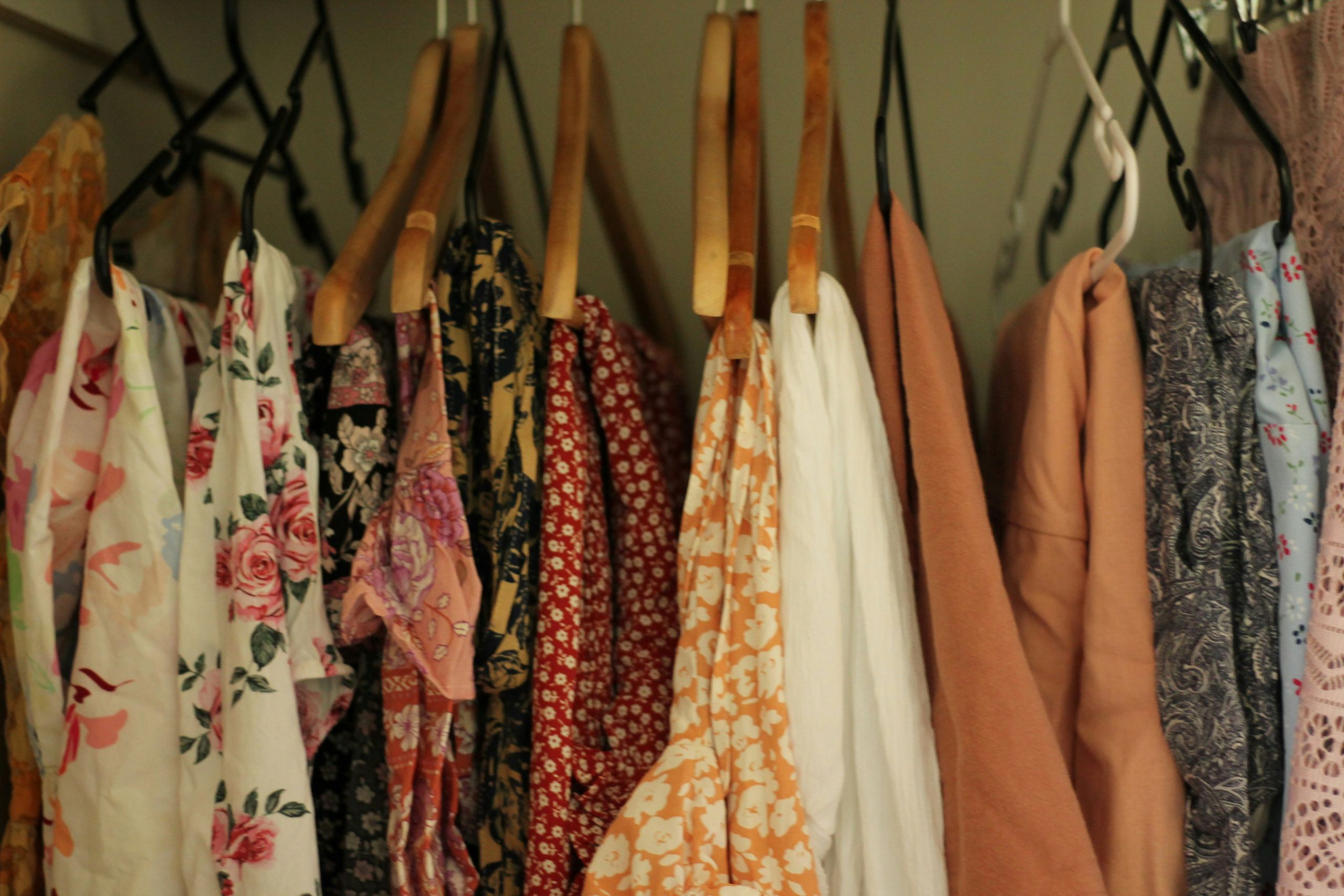 A row of dresses and tops in various floral and patterned fabrics hang on wooden hangers in a closet, featuring colors like peach, cream, pink, blue, and red.