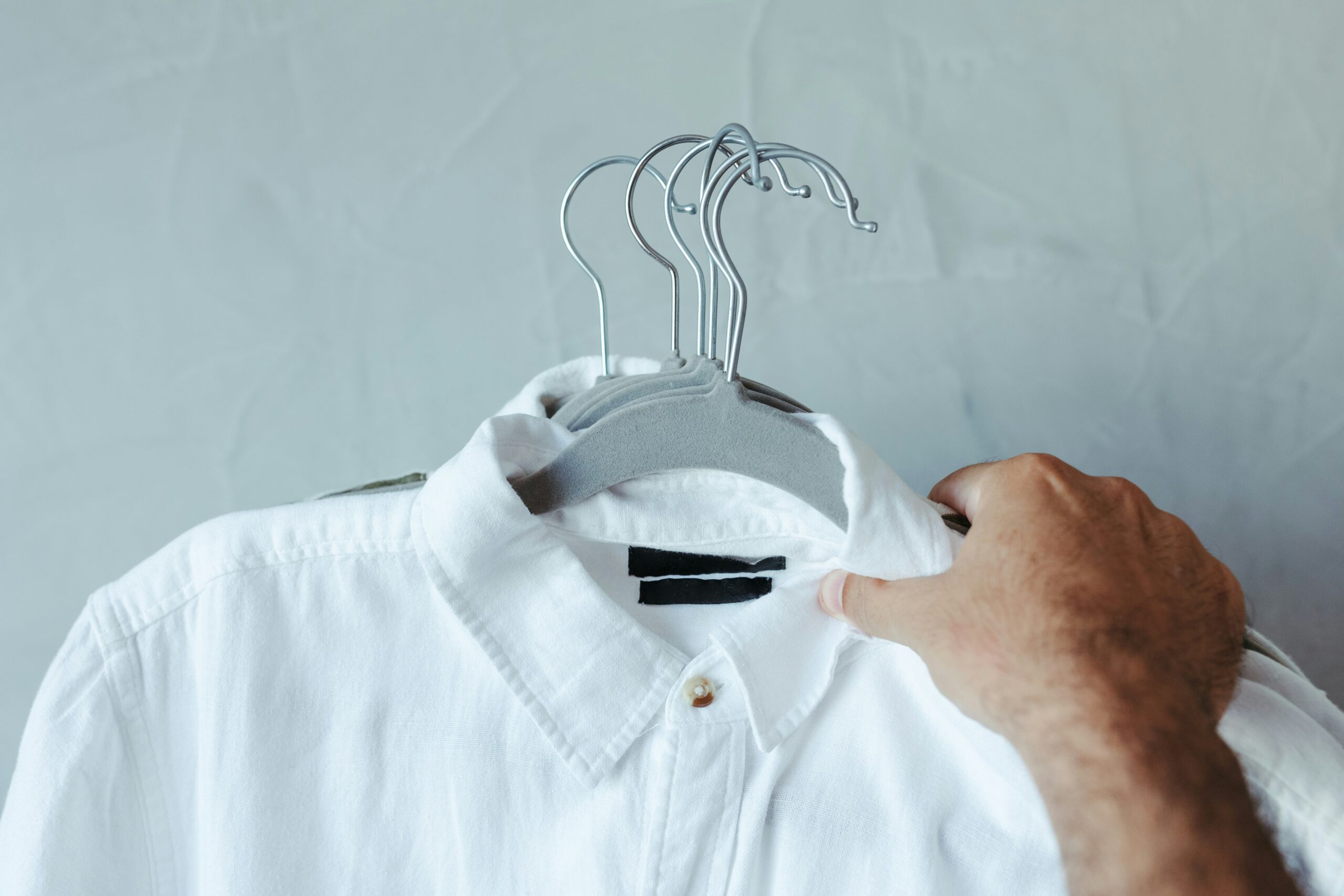 A hand holds a hanger with a white button-up shirt, with two more empty hangers stacked behind it, against a light gray textured background.