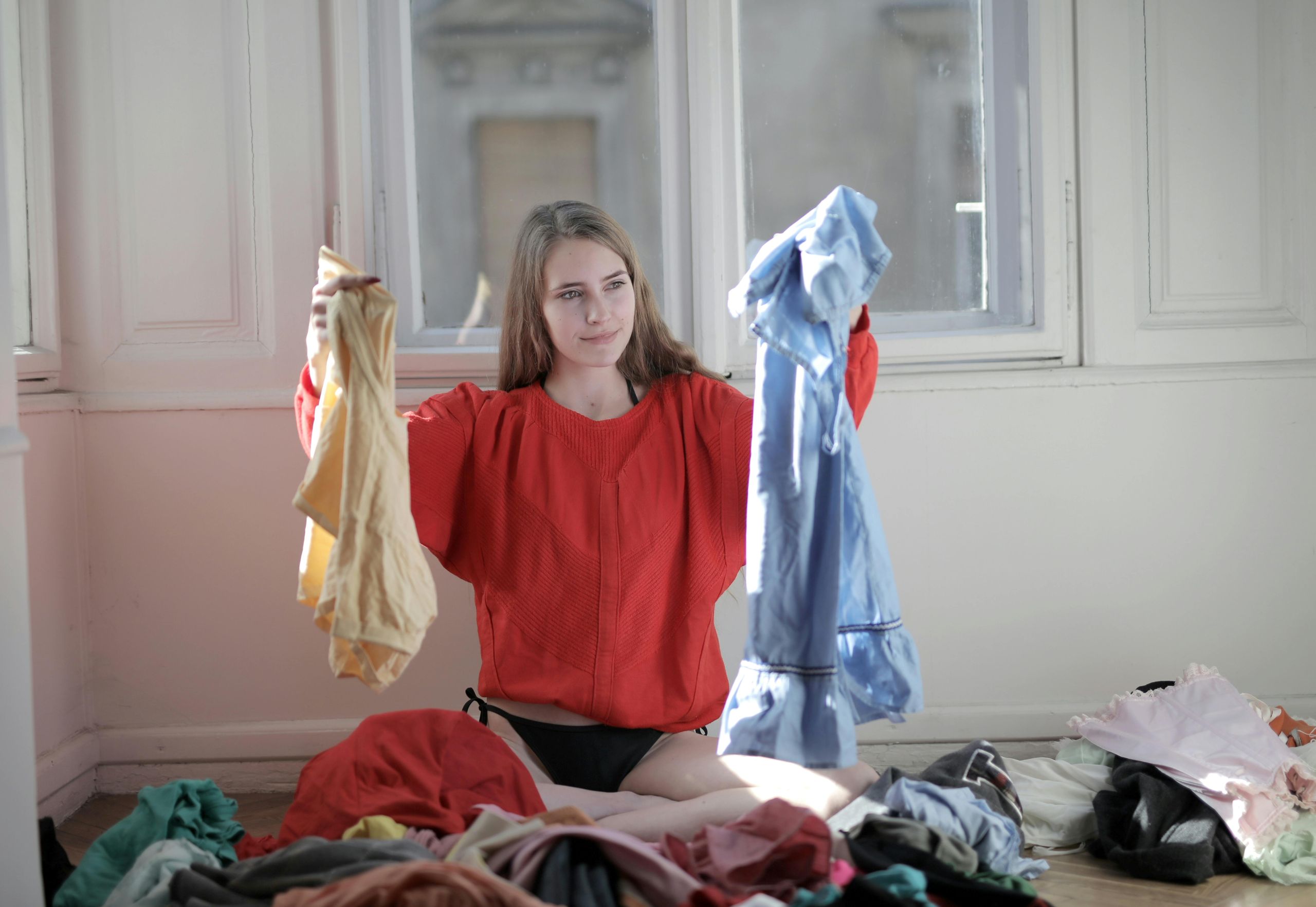 A person sits on the floor in a sunlit room, surrounded by clothes, holding up a yellow garment and a blue one, perhaps reconsidering past laundry mistakes while deciding which to wear.