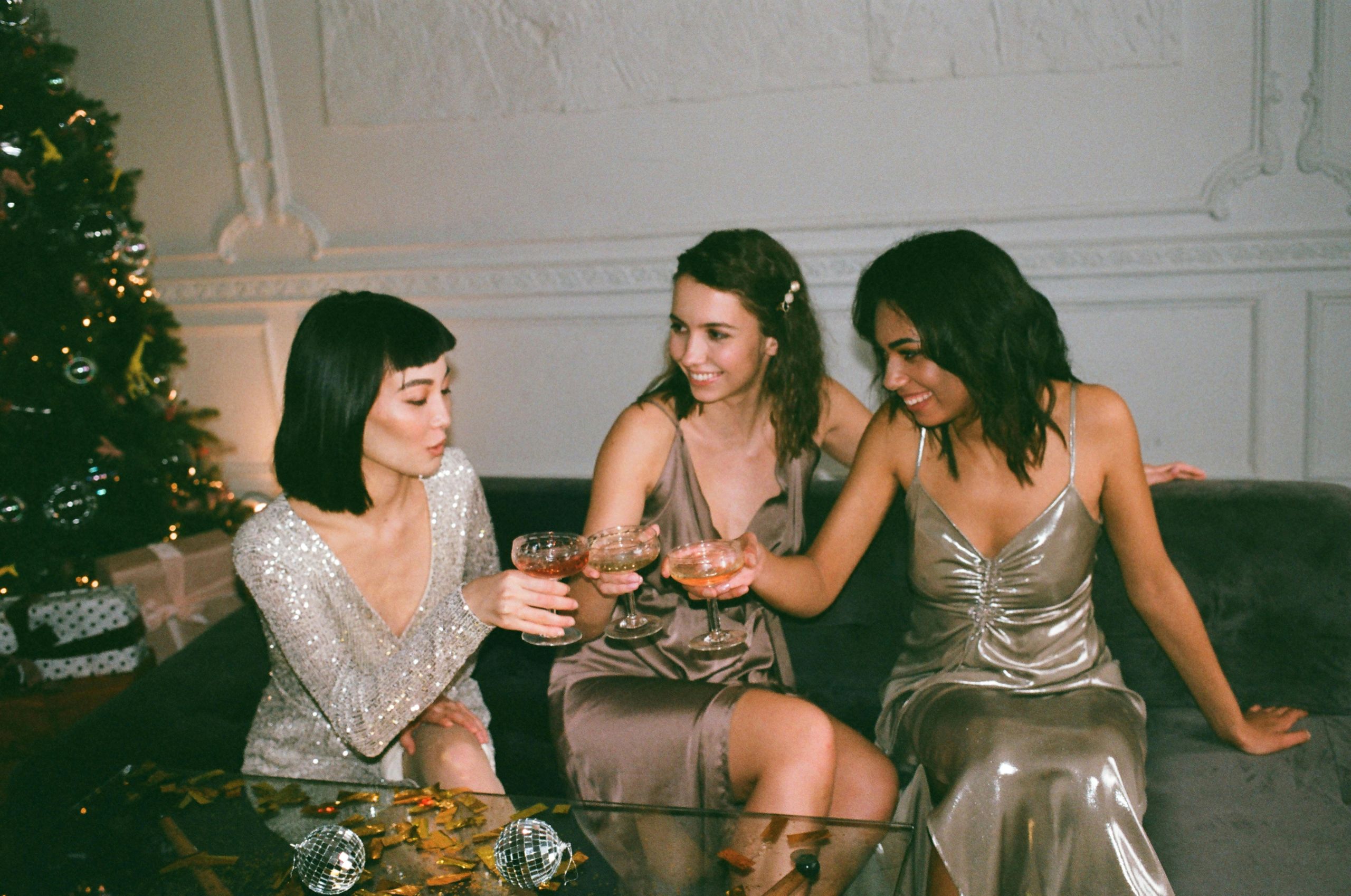 Three women in glittery dresses sit on a couch clinking glasses, smiling, and celebrating together beside a decorated Christmas tree—ready for fun, laughter, and even the occasional holiday party stains.