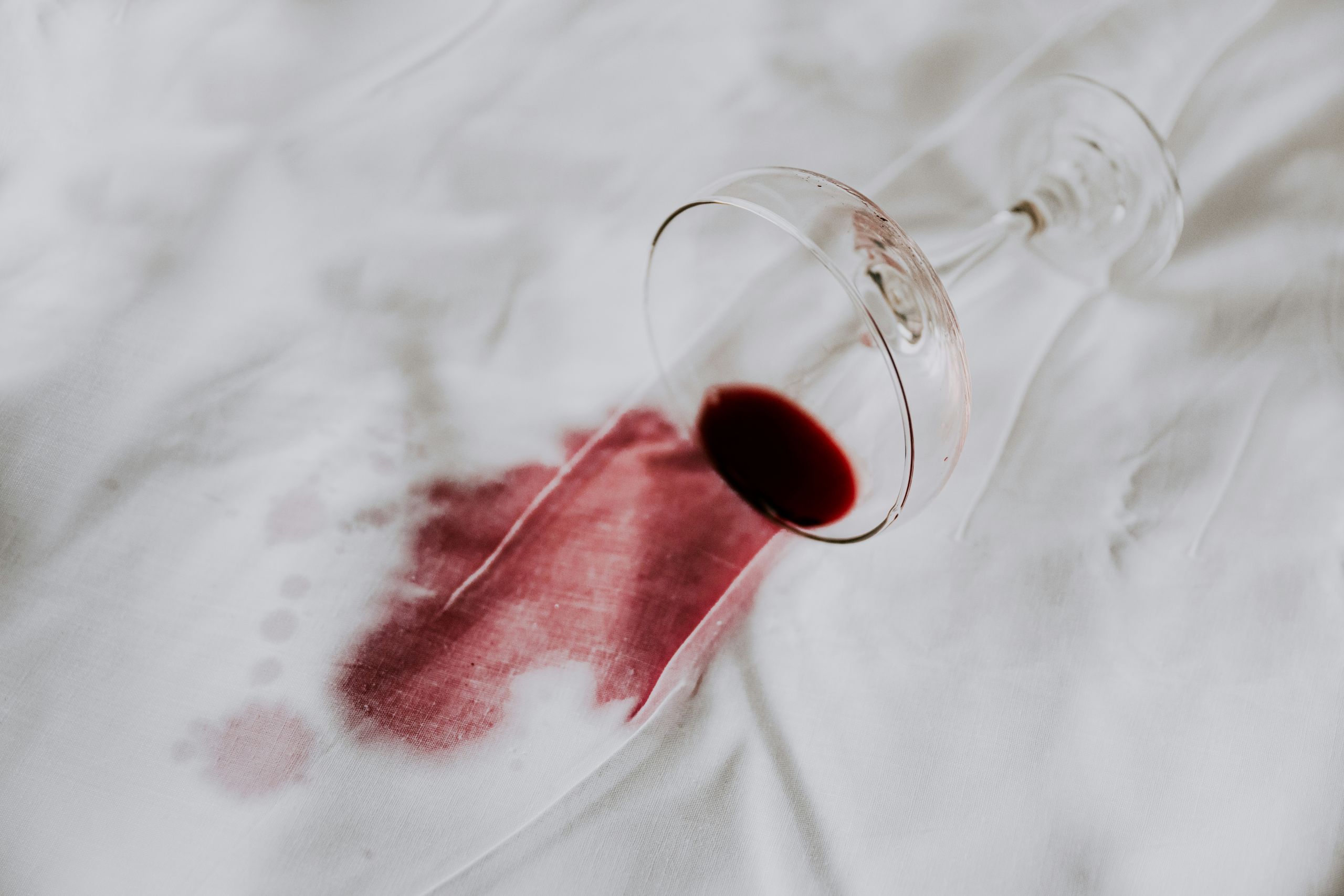 A tipped wine glass lies on a white surface, red wine spilled and creating a stubborn stain—an all-too-common scene that calls for effective wine stain removal.