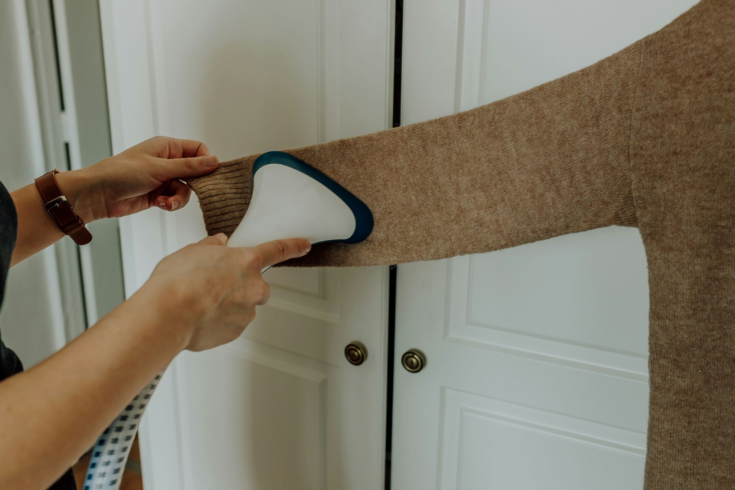 A person steams the sleeve of a brown knit sweater using a handheld garment steamer in front of white closet doors.