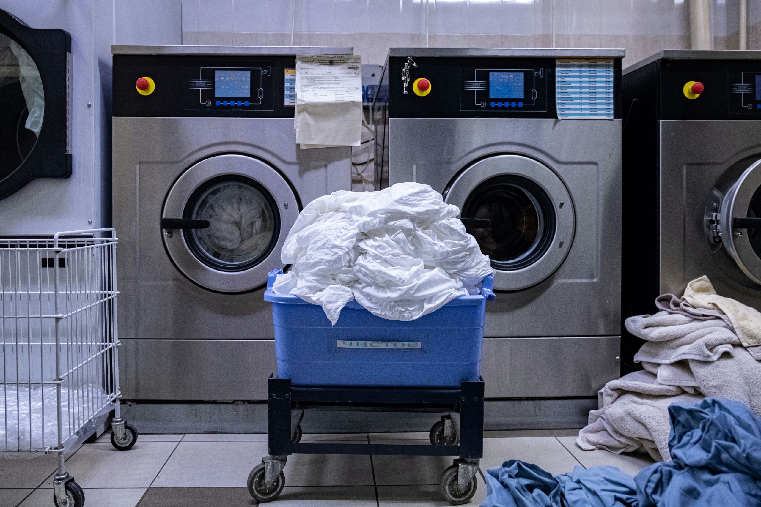 A blue cart filled with white laundry sits in front of two industrial washing machines in a laundromat, ready for a Professional Laundry Reset. Piles of laundry and an empty white basket are nearby on the tiled floor.