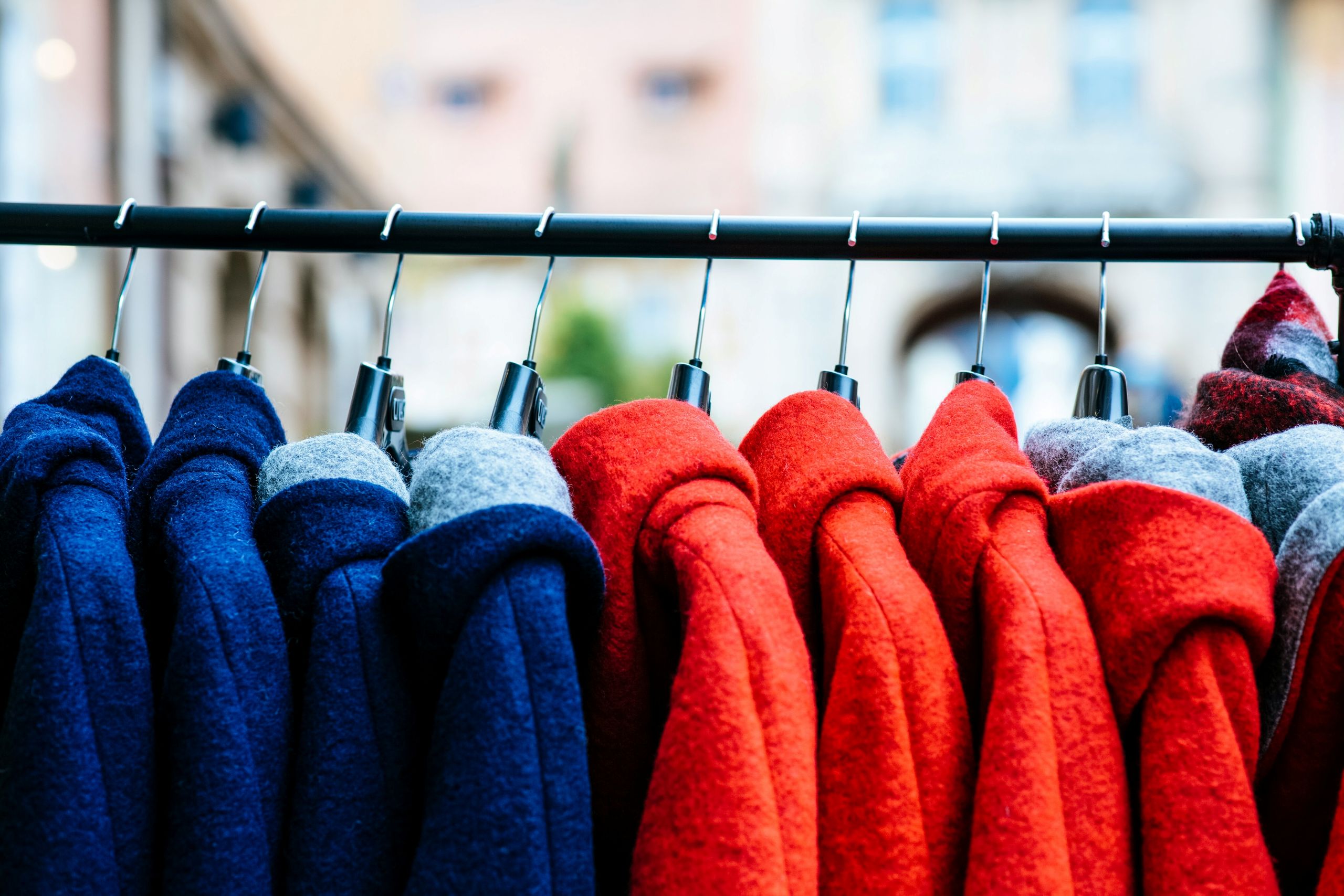 Several red and blue wool coats, exemplifying Winter Clothing Care, hang neatly on black hangers along a clothing rack, displayed outdoors with blurred buildings in the background.