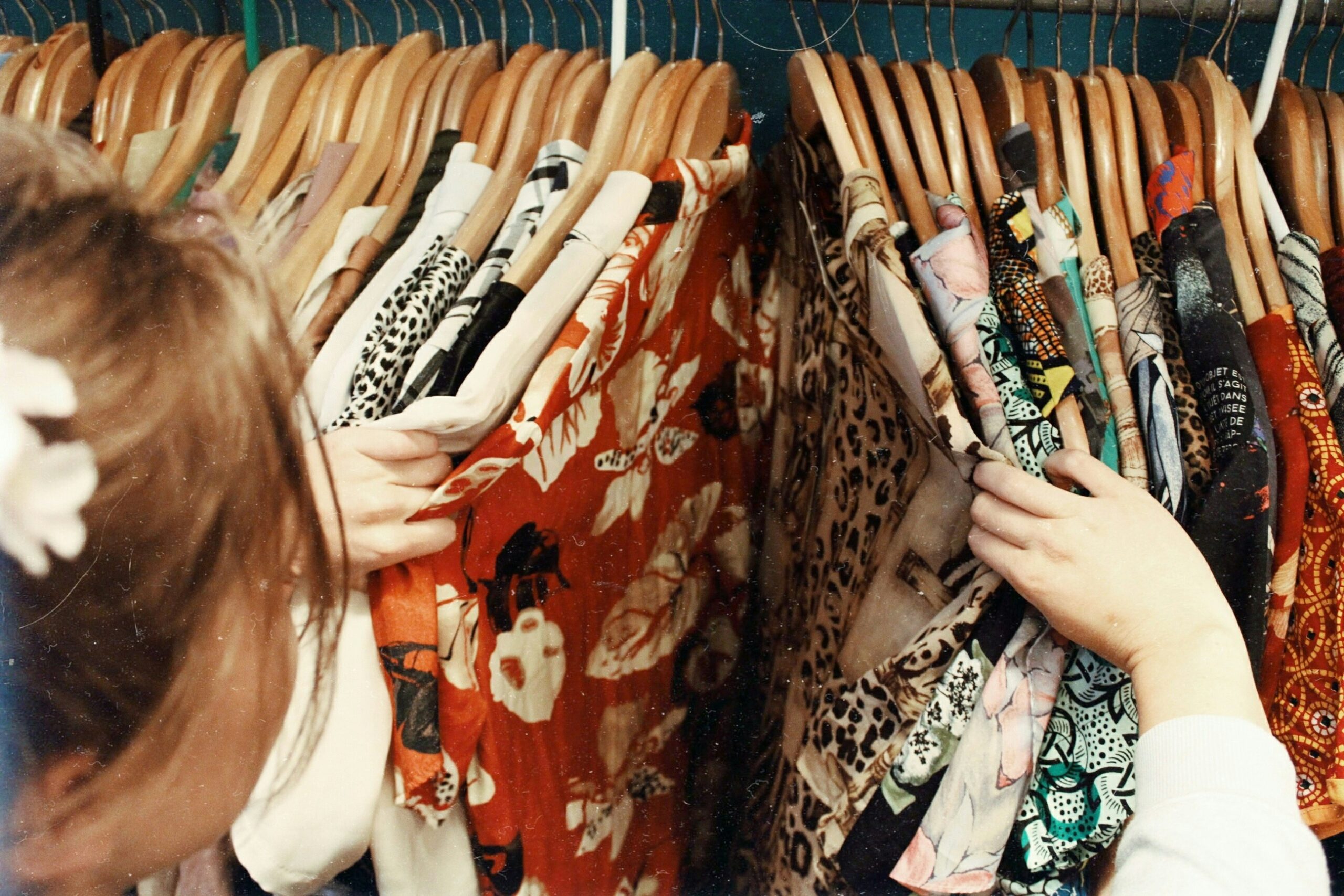 spring cleaning essentials A person browses through a rack of colorful patterned clothes on wooden hangers, inspecting different fabrics and prints during spring cleaning in a store or closet.