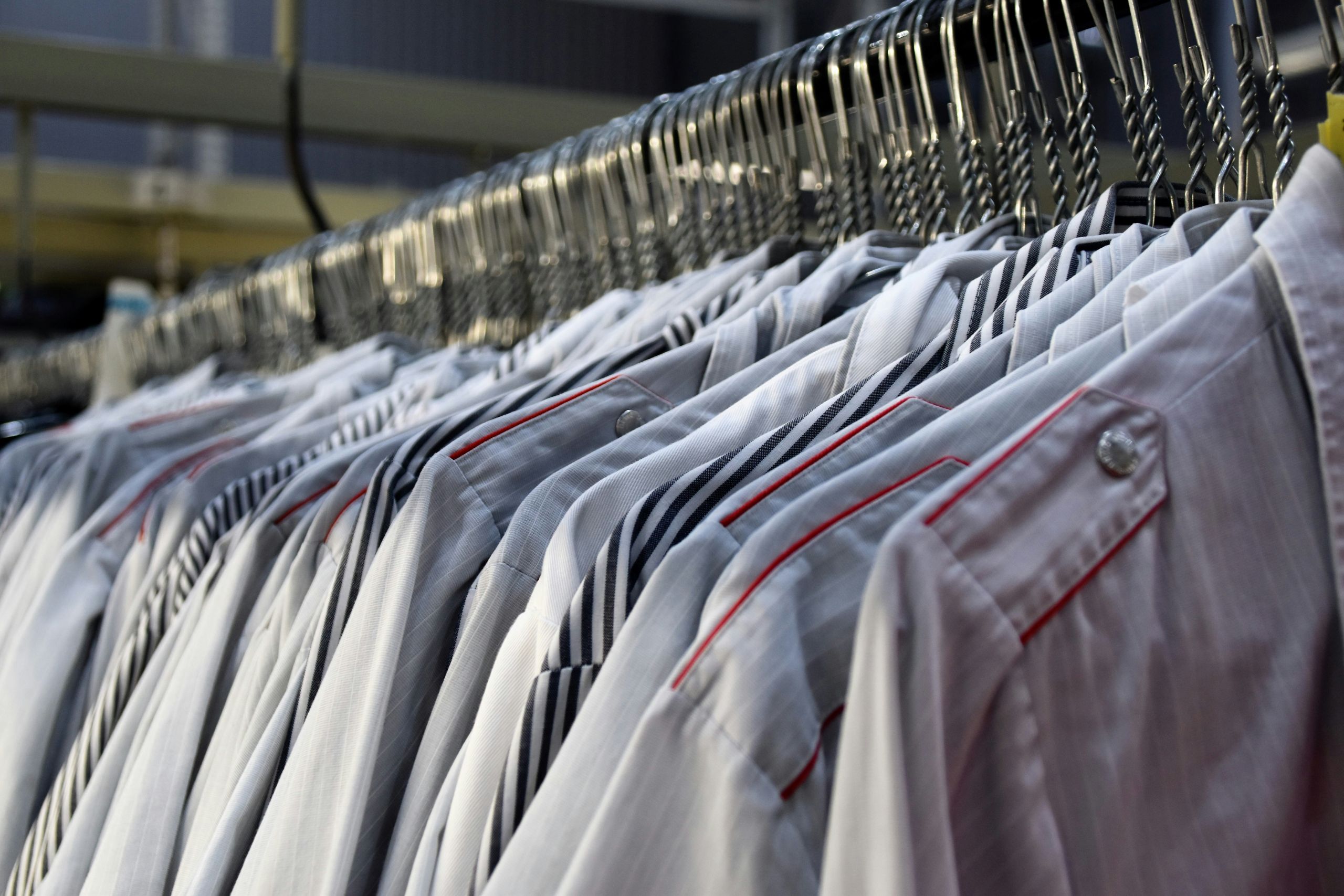 A row of neatly pressed, light-colored shirts with red piping detail on the shoulders hangs on a metal rack, showcasing the results of eco-friendly-dry-cleaning in a laundry or dry cleaning facility.