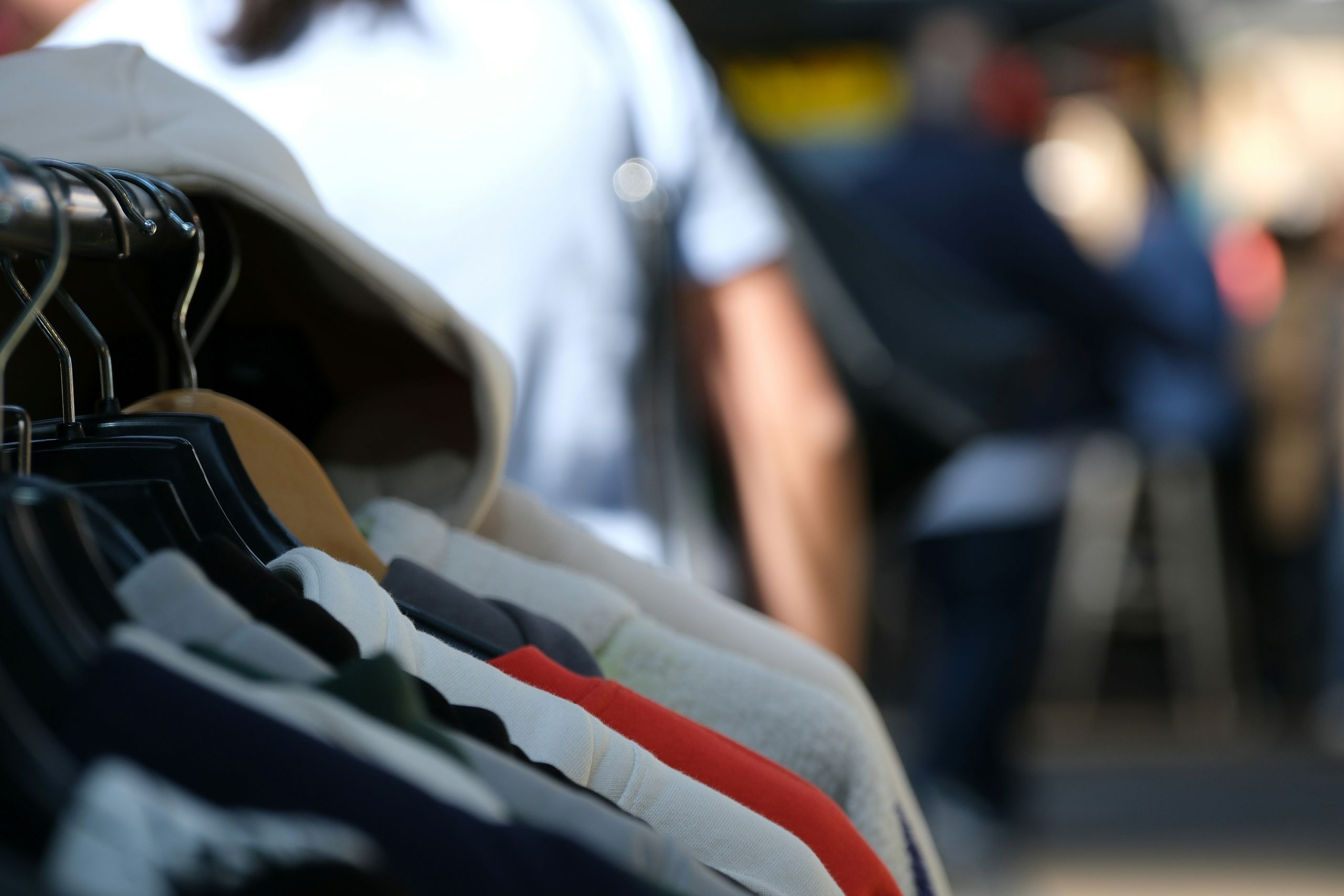 A close-up of several jackets and shirts hanging on a clothing rack outdoors, hinting at convenient dry cleaning delivery services, with blurred people and market stalls in the background.