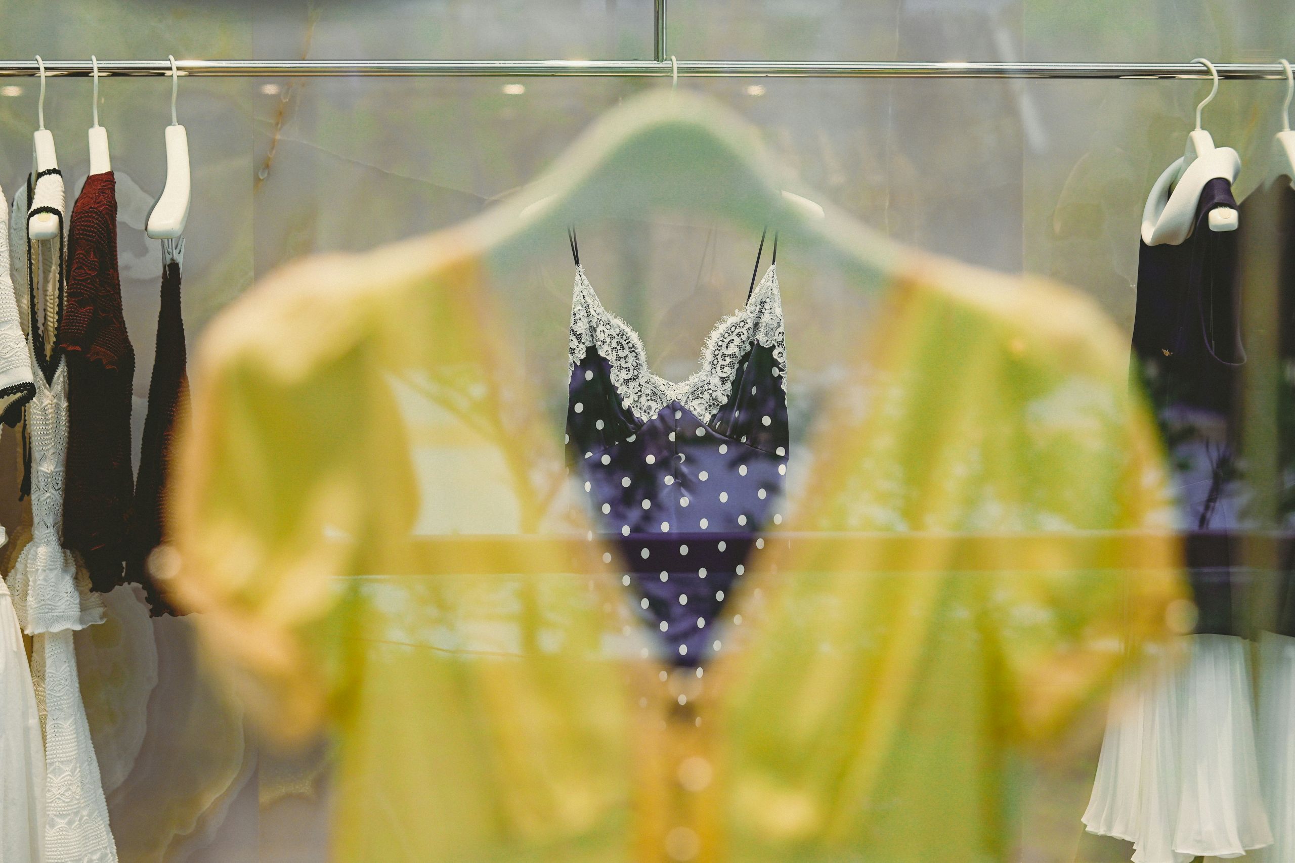 A yellow blouse on a hanger is in focus close to the camera, while a black polka-dotted, lace-trimmed camisole and other clothing items hang in the blurred background—an inviting way to keep clothes fresh.