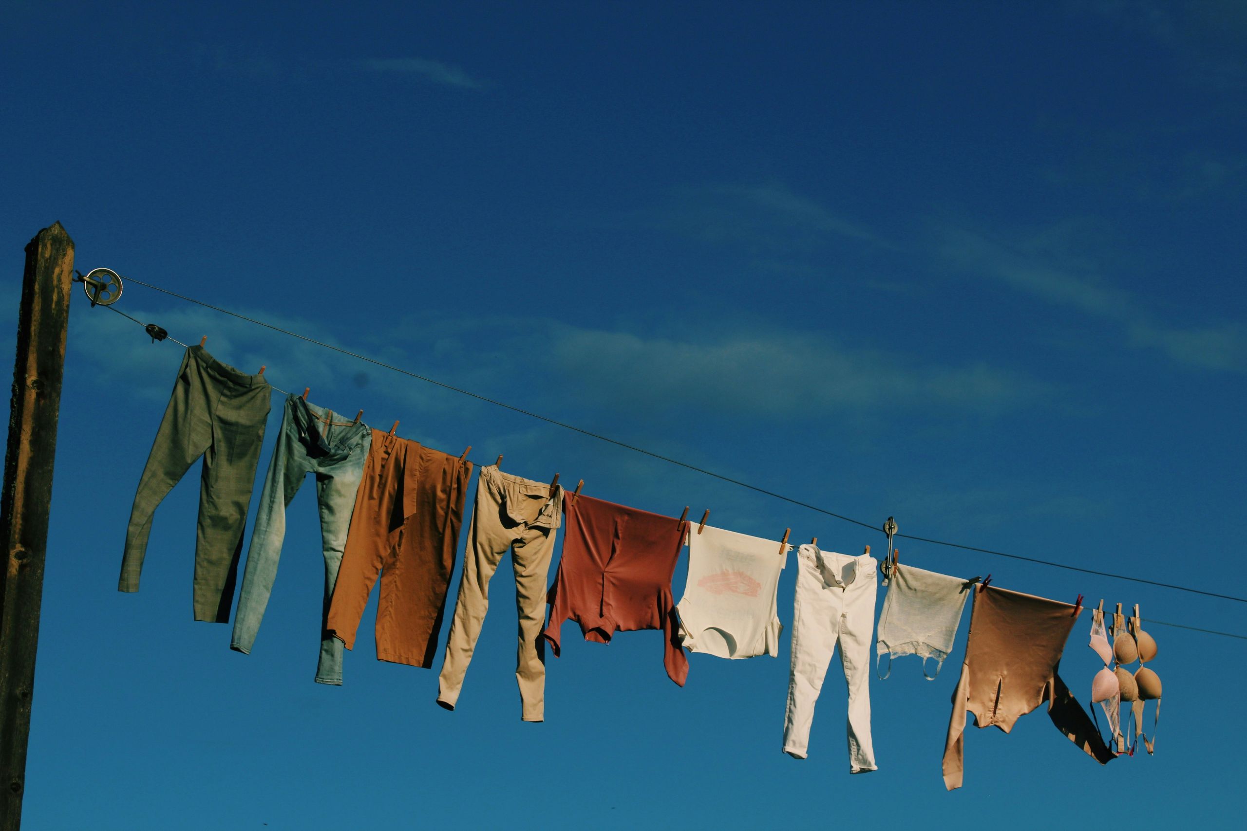 Clothes in various earth tones and a bra hang on a clothesline outdoors against a clear blue sky.