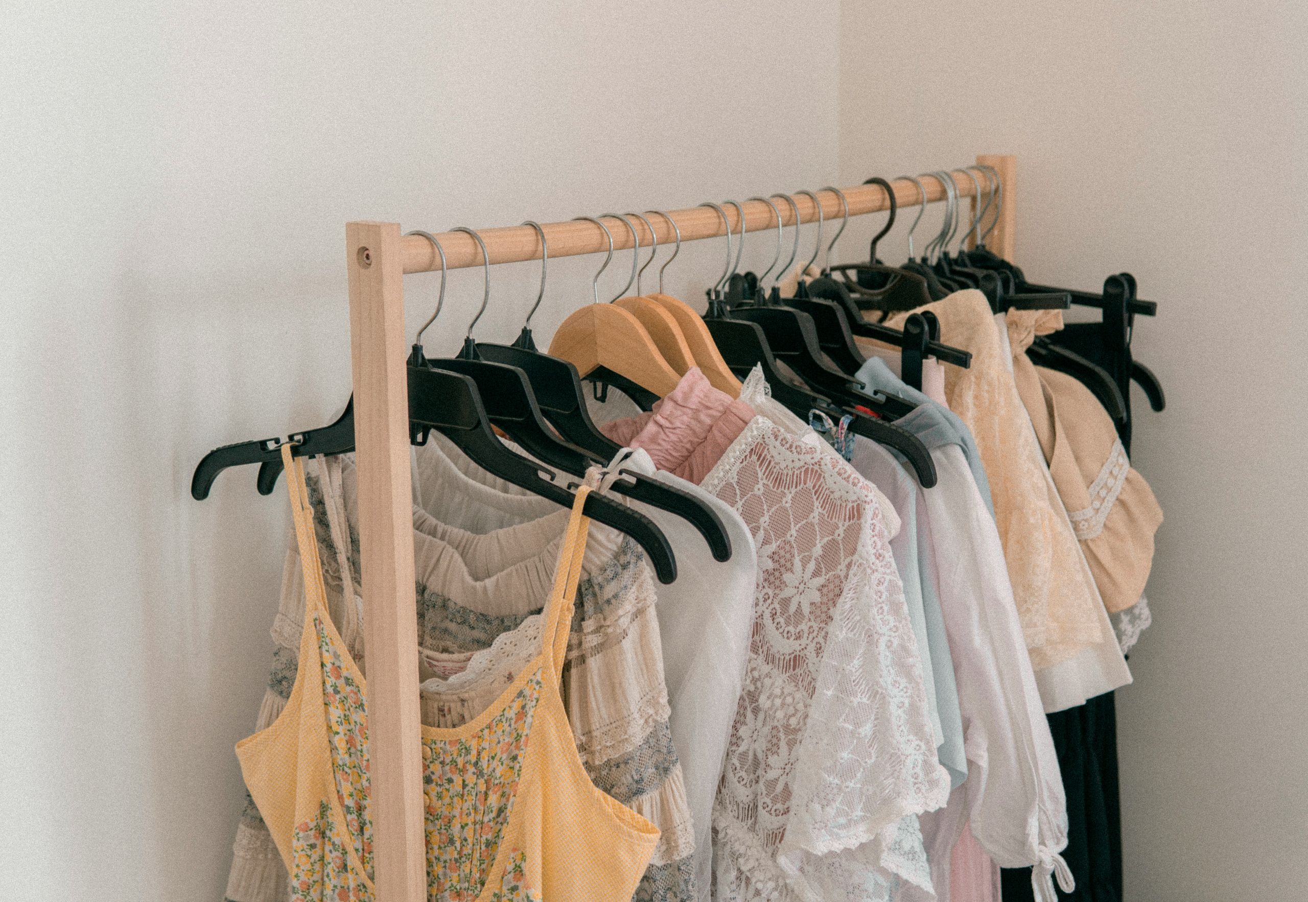 Why Professional Cleaning Helps Preserve Fabric Color A wooden clothing rack holds light-colored or clothes fading like dresses, and tops on black and wooden hangers, placed in the corner of a room with white walls.