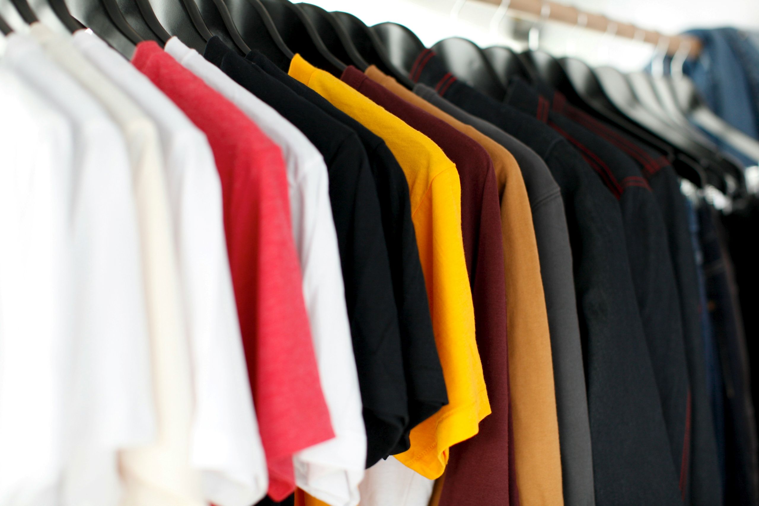 A row of assorted colored t-shirts, including white, pink, black, yellow, maroon, brown, and navy, hangs neatly on black hangers on a clothing rack.